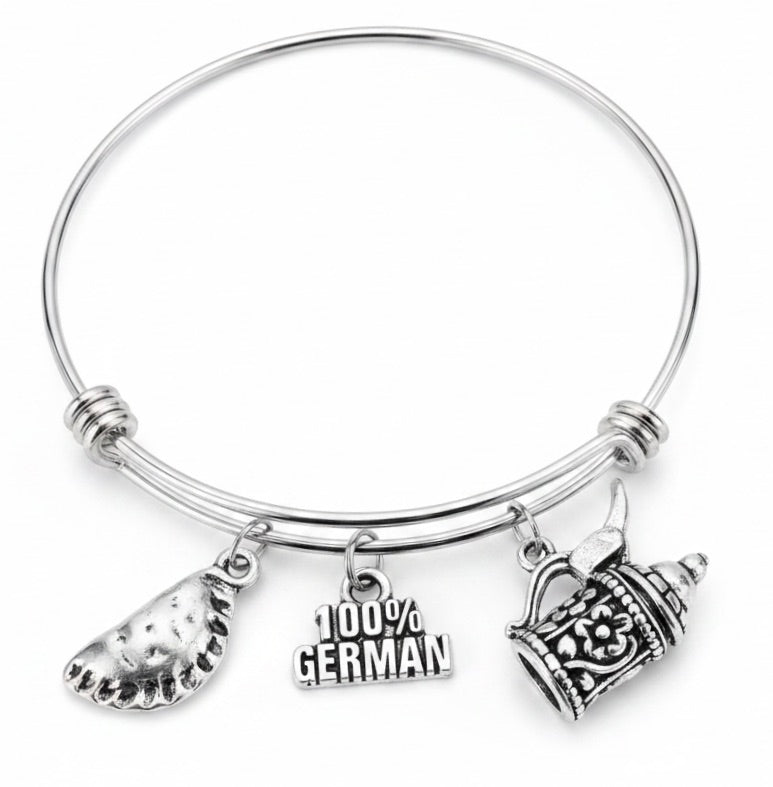 A silver-tone expandable wire bangle bracelet with three dangling charms: a crescent-shaped charm, a '100% GERMAN' text charm, and an ornate mug charm