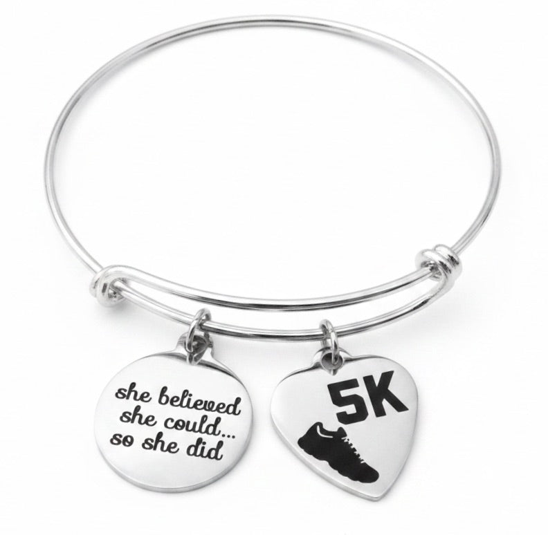 Silver expandable bangle bracelet with two charms attached via jump rings – round charm engraved 'she believed she could so she did' and heart-shaped 5K running shoe charm, studio white background