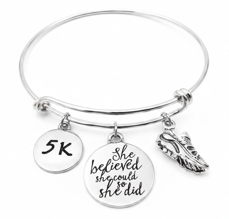 Silver expandable bangle bracelet with three charms on jump rings – small 5K disc charm, large round script charm engraved She Believed She Could So She Did, and 3D silver running shoe charm, studio white background