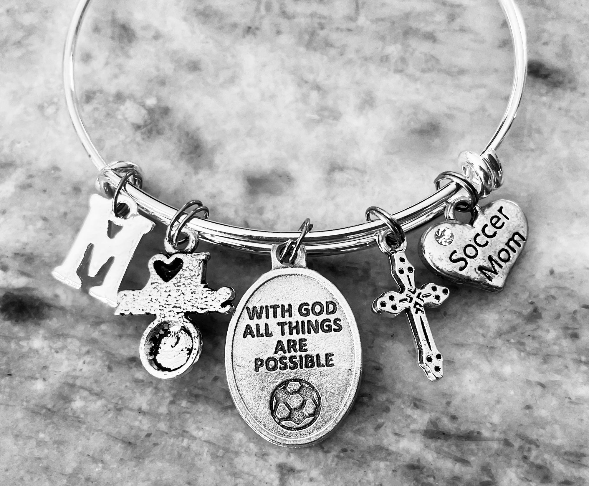silver Saint Sebastian charm bracelet showing the "With God All Things Are Possible" medal with a soccer ball, alongside cross, heart, and initial charms.