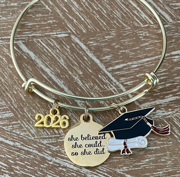 Gold expandable bangle bracelet on wood surface with three graduation charms: gold 2026 year charm, engraved disc reading she believed she could so she did, and colorful enamel graduation cap with diploma scroll and red tassel, Class of 2026 gift for her