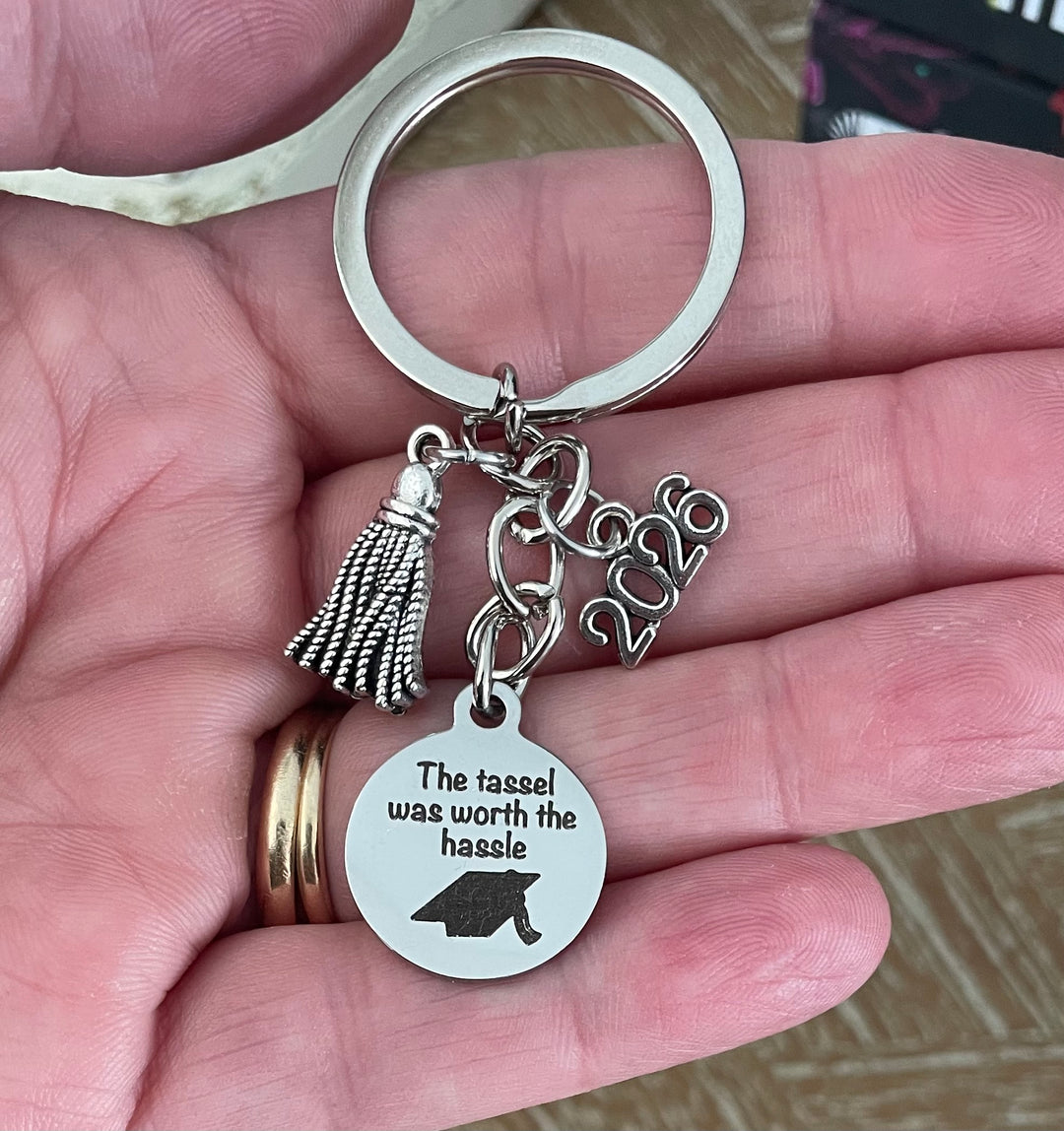 Silver graduation keychain on white background featuring tassel charm, 2026 charm, and round engraved disc with graduation cap and The tassel was worth the hassle, perfect high school or college grad gift