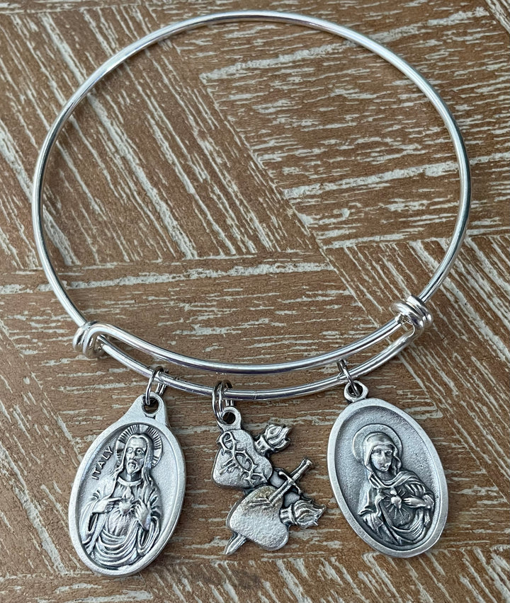 3-charm Catholic bangle bracelet on wood surface showing Sacred Heart of Jesus Italy medal, Immaculate Heart of Mary medal, and Sacred Heart rose sword charm on silver expandable wire bangle