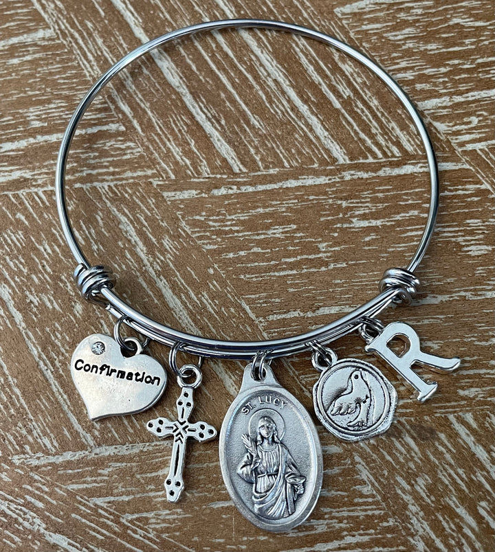 Confirmation charm bangle bracelet with St. Lucy oval saint medal, cross charm, dove medallion, Confirmation heart, and letter initial charm 