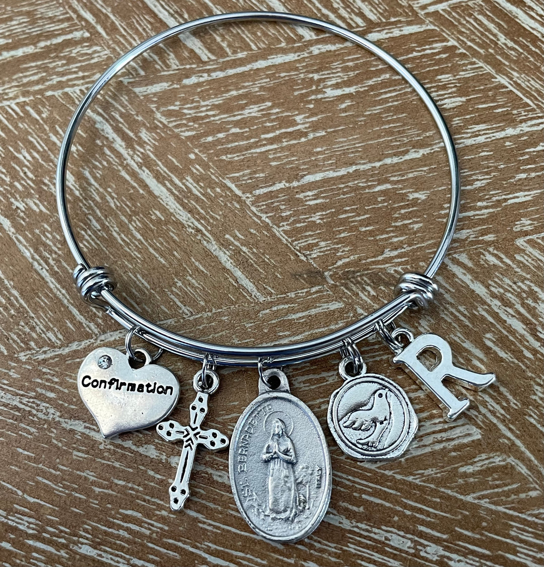 Confirmation charm bangle bracelet featuring St. Bernadette saint medal, decorative cross charm, Confirmation heart, Holy Spirit dove medallion, and personalized silver initial charm