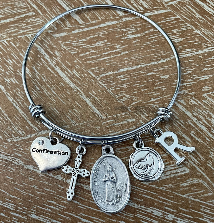 Confirmation charm bangle bracelet featuring St. Bernadette saint medal, decorative cross charm, Confirmation heart, Holy Spirit dove medallion, and personalized silver initial charm