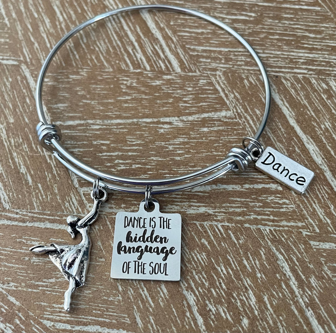 Silver-plated expandable charm bangle bracelet with ballerina dancer charm, engraved "Dance is the Hidden Language of the Soul" square charm, and Dance tag charm, displayed on rustic