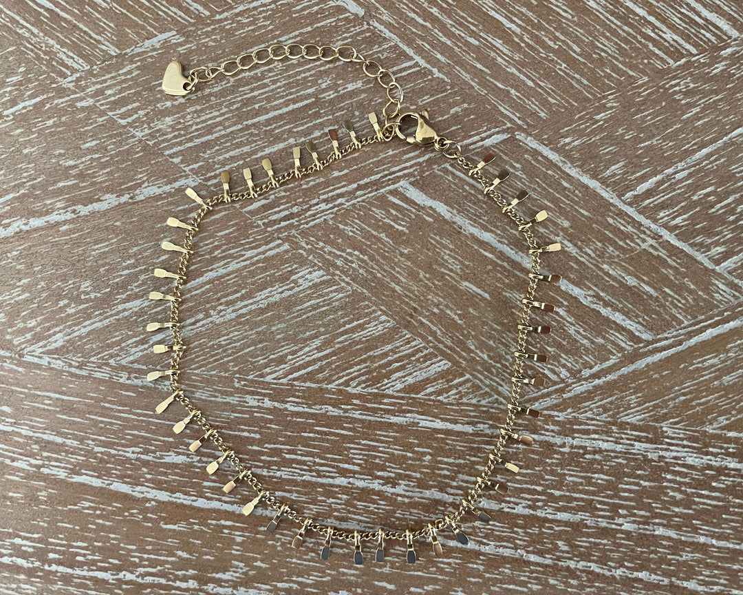 A gold-toned anklet with multiple small flat metal spikes and dangling teardrop charms, laid flat on a brown wood-textured surface.