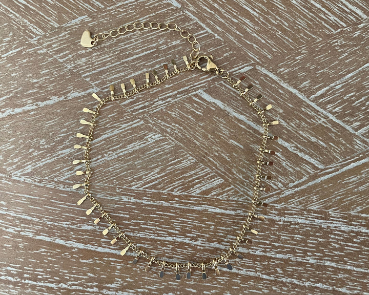 A gold-toned anklet with multiple small flat metal spikes and dangling teardrop charms, laid flat on a brown wood-textured surface.