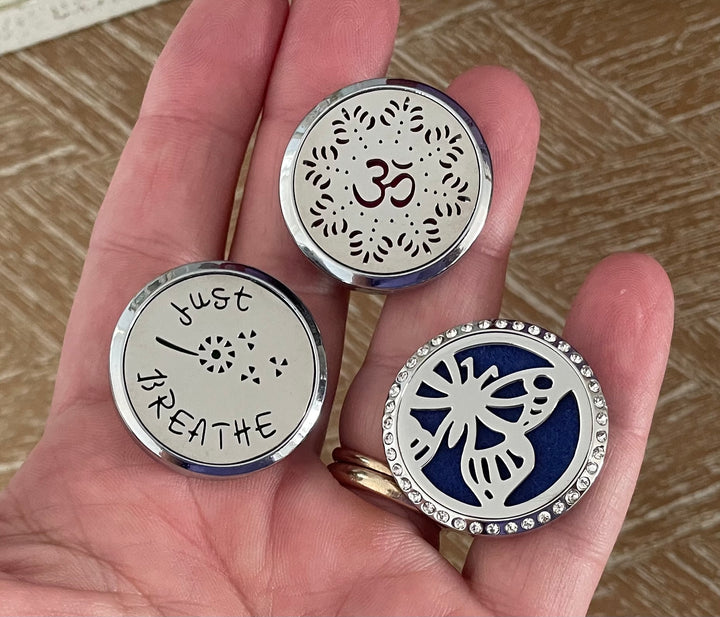 Three stainless steel essential oil car diffuser lockets on wood surface — Om symbol, butterfly crystal rhinestone, and Just Breathe designs with vent clips