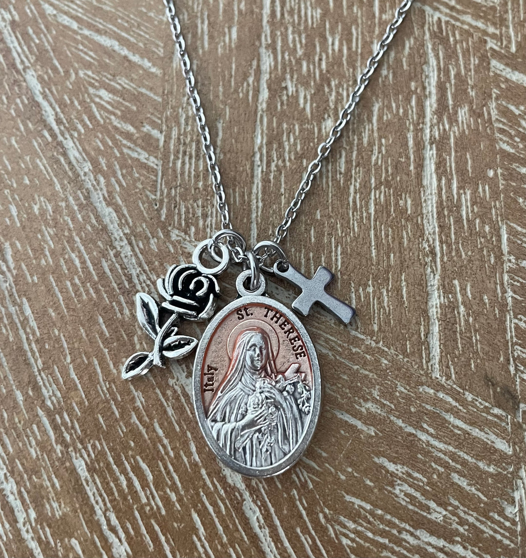 Silver chain necklace on white background featuring an oval Italian St. Therese of Lisieux religious medal with rose-gold enamel center and bas-relief image of St. Therese holding flowers, a silver rose bud charm with stem and leaves, and a small silver Latin cross charm
