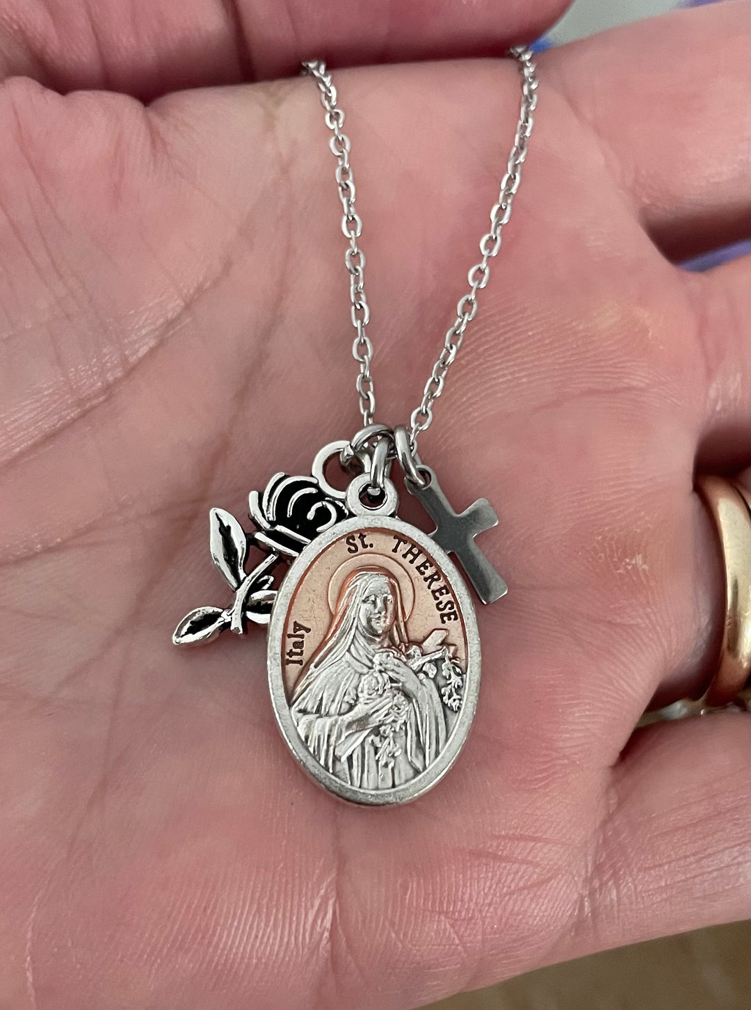 A silver St. Teresa of Calcuta oval pendant necklace with a rose charm and a small cross charm, held in a person&