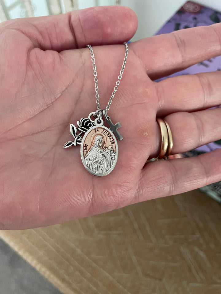 St. Therese of Lisieux Italian oval medal charm with rose-gold enamel center, raised silver border, and accompanying silver rose bud and cross charms on stainless steel chain