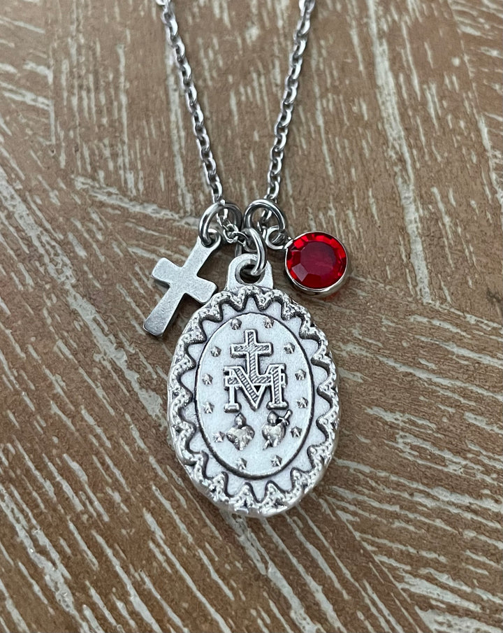Silver-tone necklace with a monogrammed oval medallion, a small cross charm, and a round gemstone, displayed on a textured brown wood background.