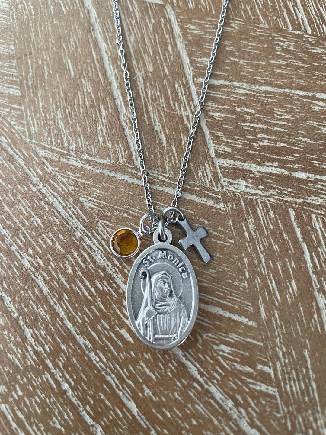 A silver St. Monica medal necklace with a small amber gemstone and a silver cross charm, displayed on a textured brown wooden surface.