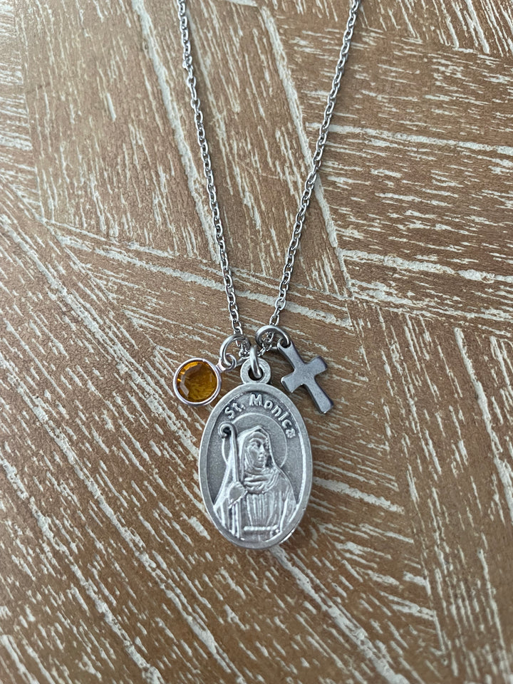 A silver St. Monica medal necklace with a small amber gemstone and a silver cross charm, displayed on a textured brown wooden surface.