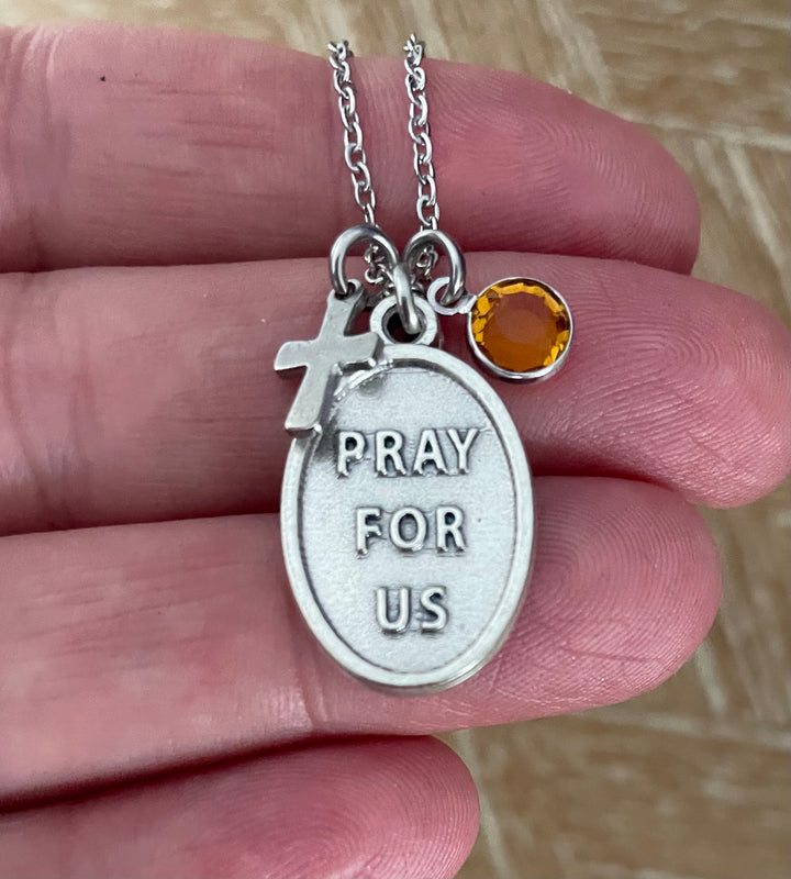 A close-up of a silver necklace held in a hand, featuring an oval pendant with the words &