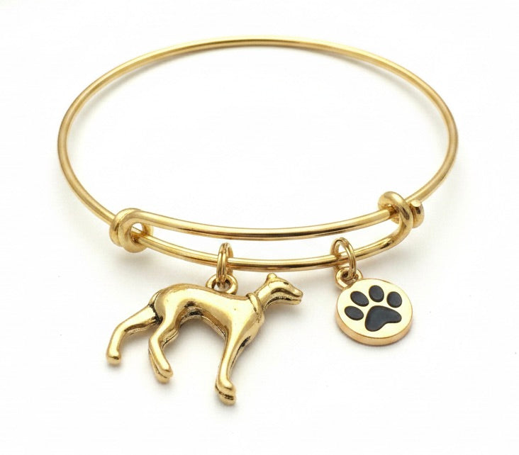 Gold bangle bracelet with dangling charm of a greyhound dog and a round charm featuring a black paw print, on a white background.