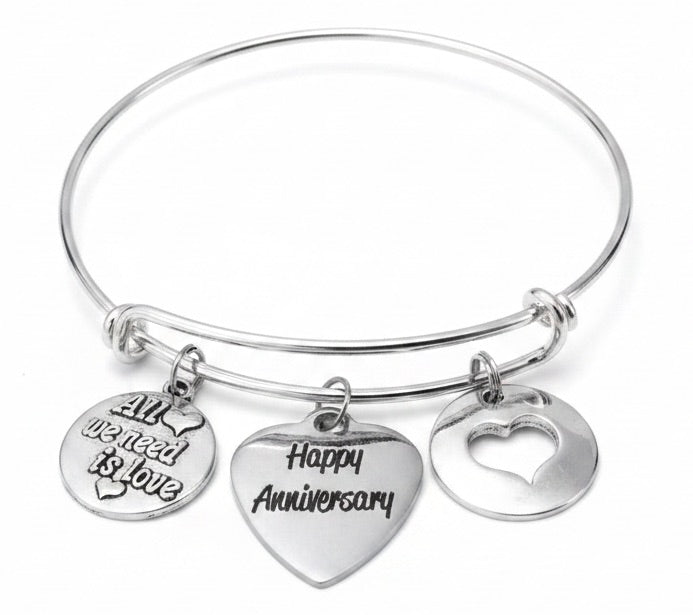 Silver expandable bangle bracelet with three charms: All We Need is Love engraved disc, Happy Anniversary heart charm, and heart cutout disc — romantic anniversary gift for wife