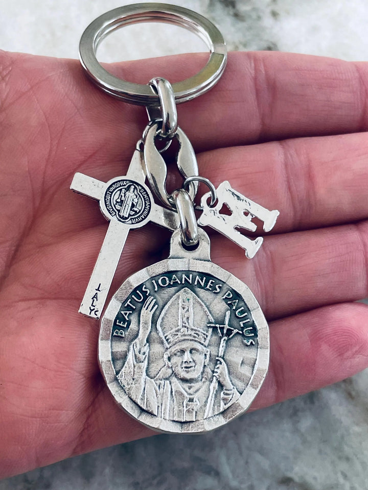 A silver-toned metal keychain with multiple charms, including a round saint medallion, a cross, and a small letter charm,