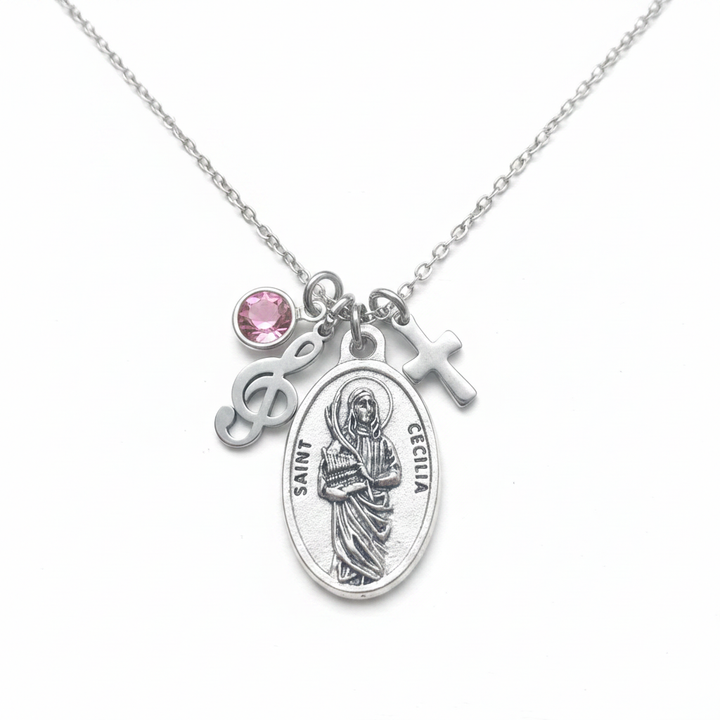 Silver Saint Cecilia oval medal necklace with treble clef music charm, cross charm, and pink faceted crystal birthstone on stainless steel cable chain, white studio background