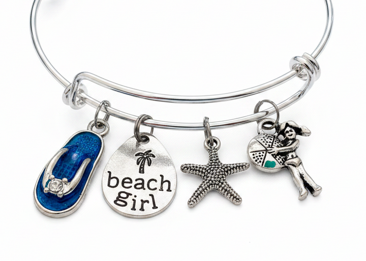Silver expandable bangle bracelet with four beach-themed charms: blue enamel flip flop with crystal accent, teardrop beach girl tag with palm tree, silver starfish, and beach girl figure holding teal enamel beach ball — adjustable stackable gift bracelet for women