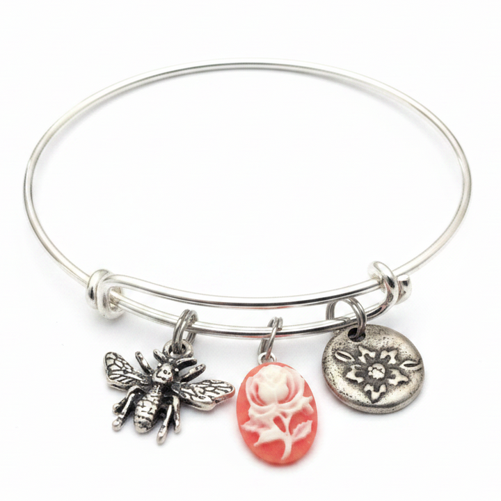 Silver expandable bangle bracelet with three charms: detailed silver honeybee with textured wings, round embossed silver flower disc, and pink oval cameo with white rose relief carving — adjustable stackable vintage style gift bracelet for women