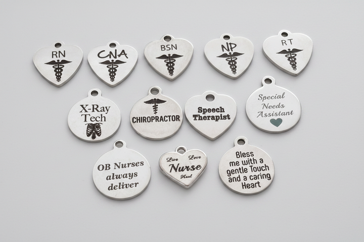 Collection of silver stainless steel engraved charms for healthcare professionals including RN, CNA, BSN, NP, RT, X-Ray Tech, Chiropractor, Speech Therapist, Special Needs Assistant, and nurse-themed sentiment charms, arranged on a light background with studio lighting