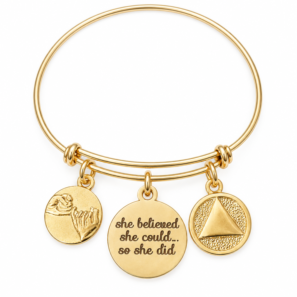 Gold bangle bracelet with a pinky swear charm, a She Believed She Could So She Did engraved charm, and an AA recovery symbol medallion charm, shown on a warm-white studio background