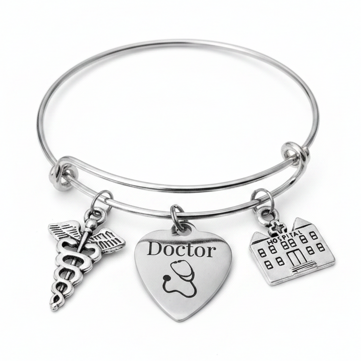 Silver stainless steel bangle charm bracelet featuring a caduceus medical symbol charm, a heart charm engraved with Doctor and stethoscope, and a hospital building charm — gift-ready jewelry for doctors and medical professionals