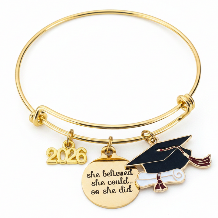 Gold graduation charm bangle bracelet on white background featuring 2026 charm, she believed she could engraved disc, and enamel graduation cap with diploma, professional product photo