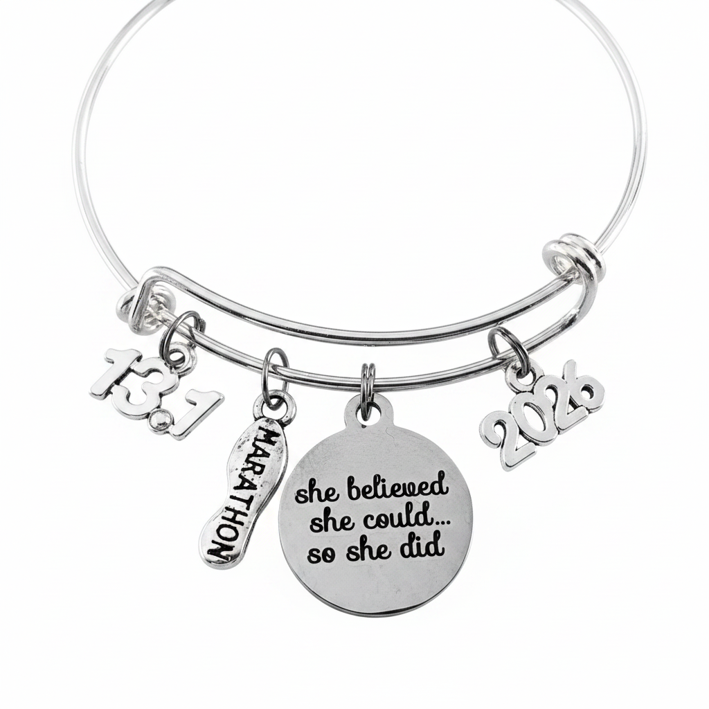 Silver expandable bangle bracelet with four charms on jump rings – 13.1 number charm, Marathon running shoe charm, large round disc engraved She Believed She Could So She Did in script, and 2026 year charm, studio white background