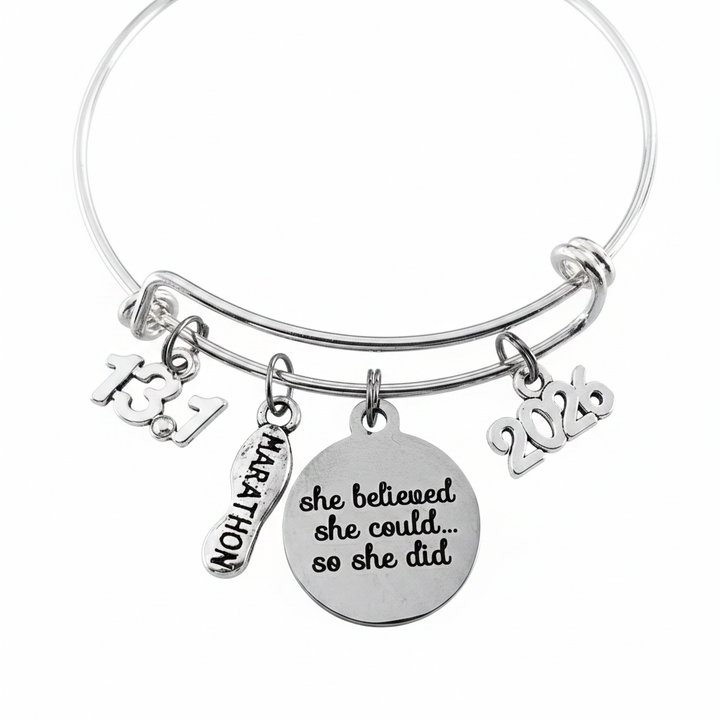 Silver expandable bangle bracelet with four charms on jump rings – 13.1 number charm, Marathon running shoe charm, large round disc engraved She Believed She Could So She Did in script, and 2026 year charm, studio white background