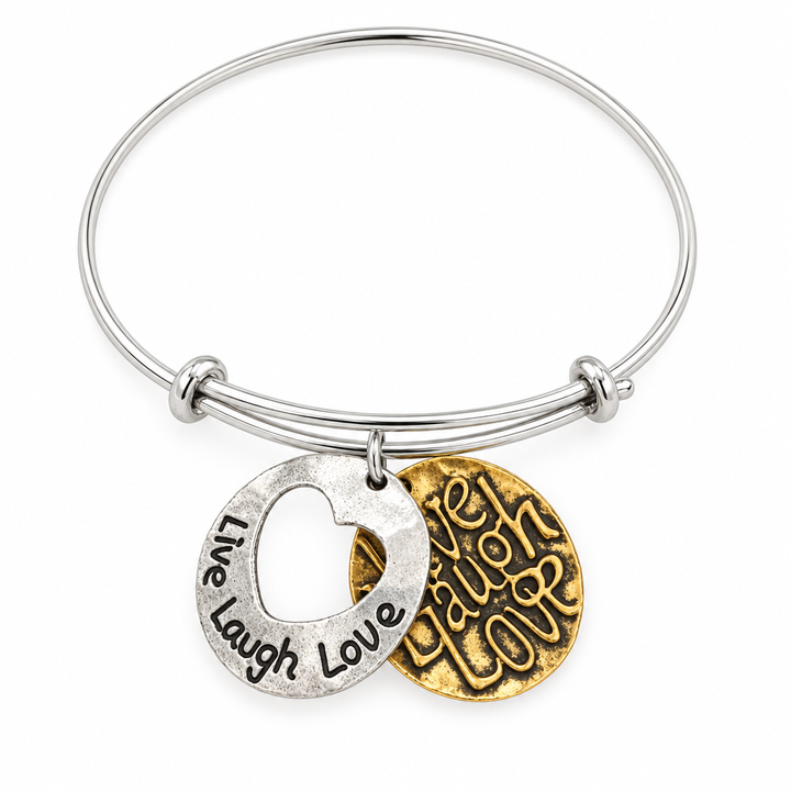 Silver bangle bracelet with two Live Laugh Love charms, one silver with a heart cutout and one gold hammered-texture, shown on a warm-white studio background
