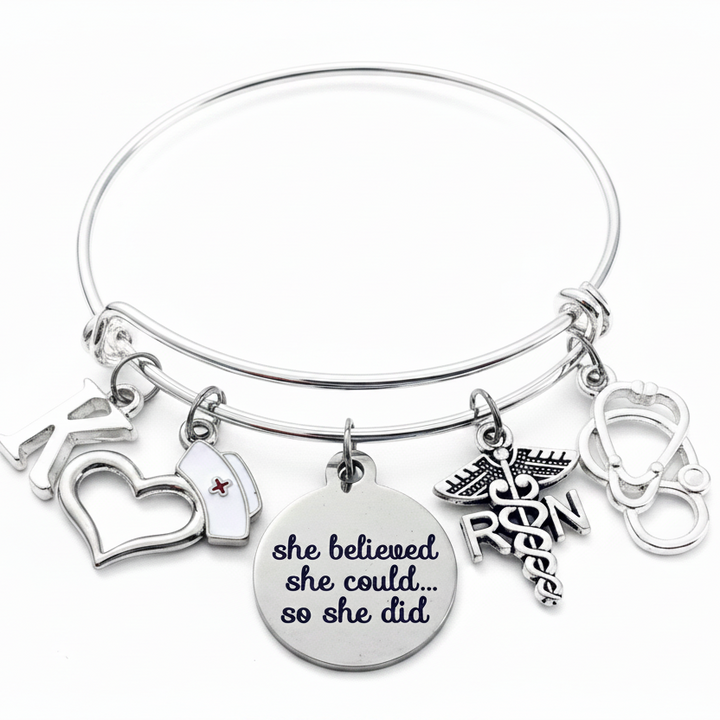 Silver expandable bangle bracelet with nurse-themed charms including RN caduceus, stethoscope, nurse cap, open heart, and inspirational disc charm reading she believed she could so she did, on white background