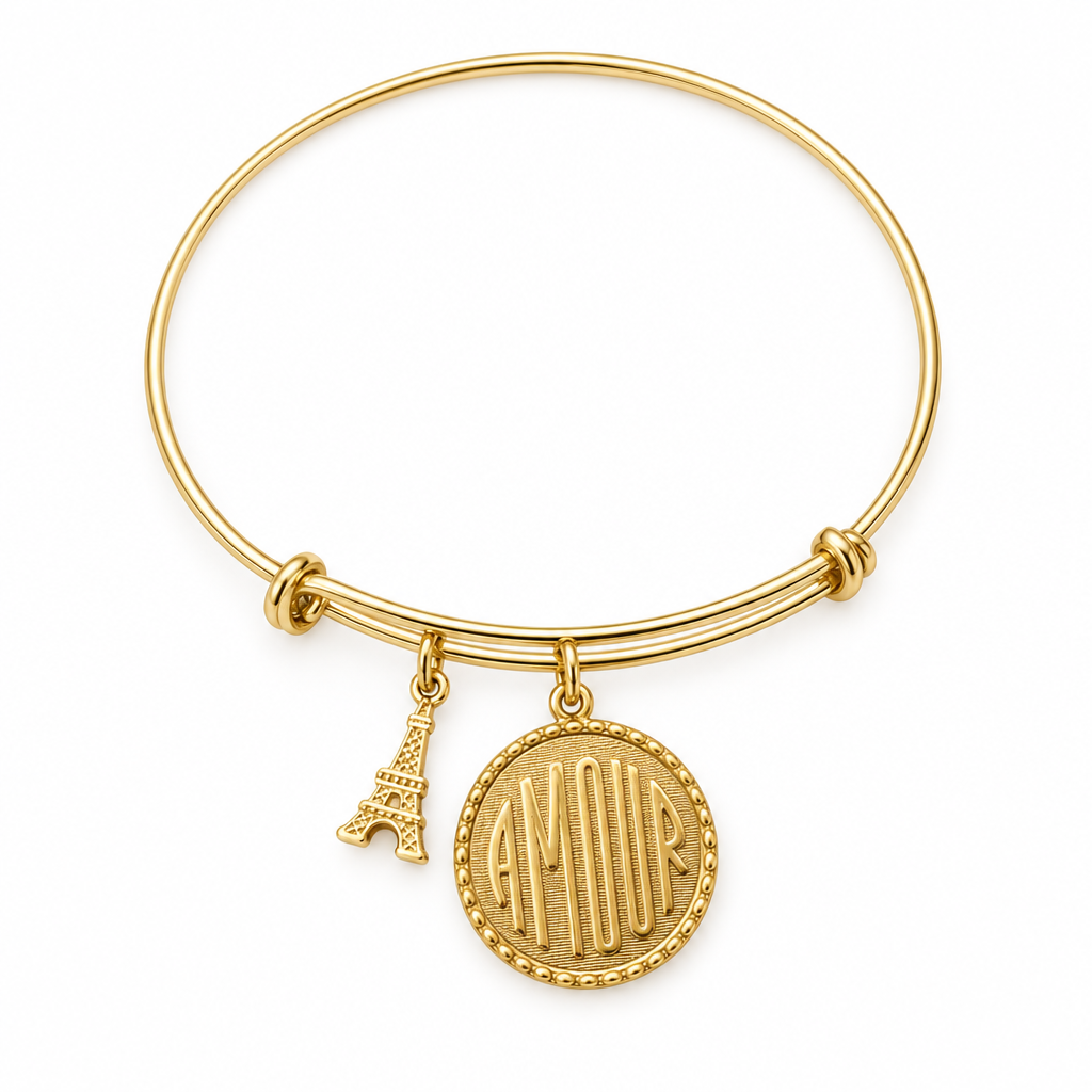 Gold bangle bracelet with an AMOUR coin charm and a 3D Eiffel Tower charm, shown on a warm-white studio background