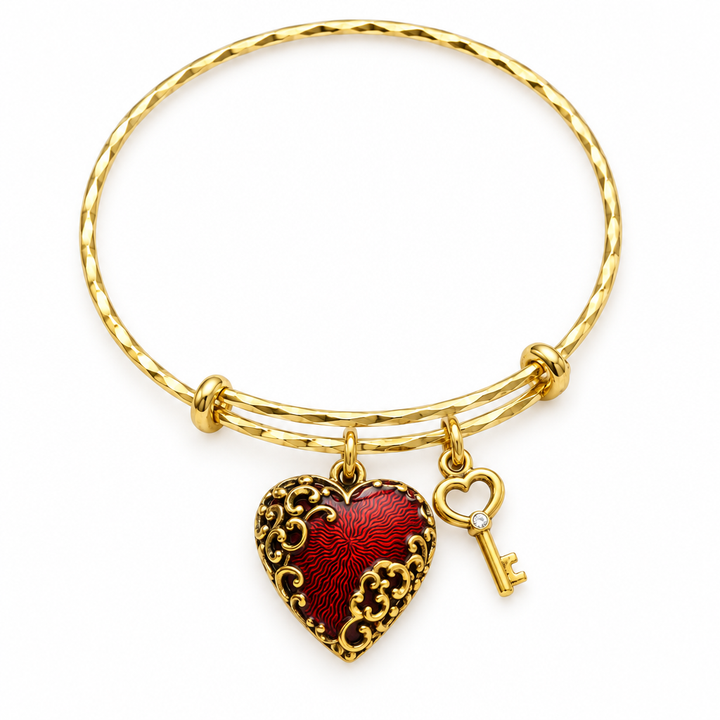 Gold twisted bangle bracelet with a red enamel filigree heart charm and a gold key charm, shown on a warm-white studio background