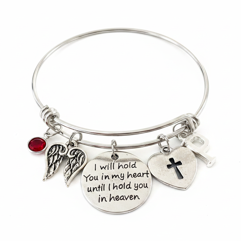 Silver expandable bangle bracelet with memorial charms — red crystal birthstone, angel wings, I will hold you in my heart sentiment disc, cross heart, and initial charm — on white background, handcrafted sympathy gift jewelry by Jules Obsession