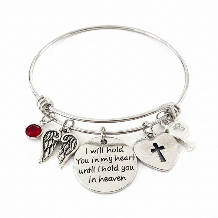 Silver expandable bangle bracelet with memorial charms — red crystal birthstone, angel wings, I will hold you in my heart sentiment disc, cross heart, and initial charm — on white background, handcrafted sympathy gift jewelry by Jules Obsession