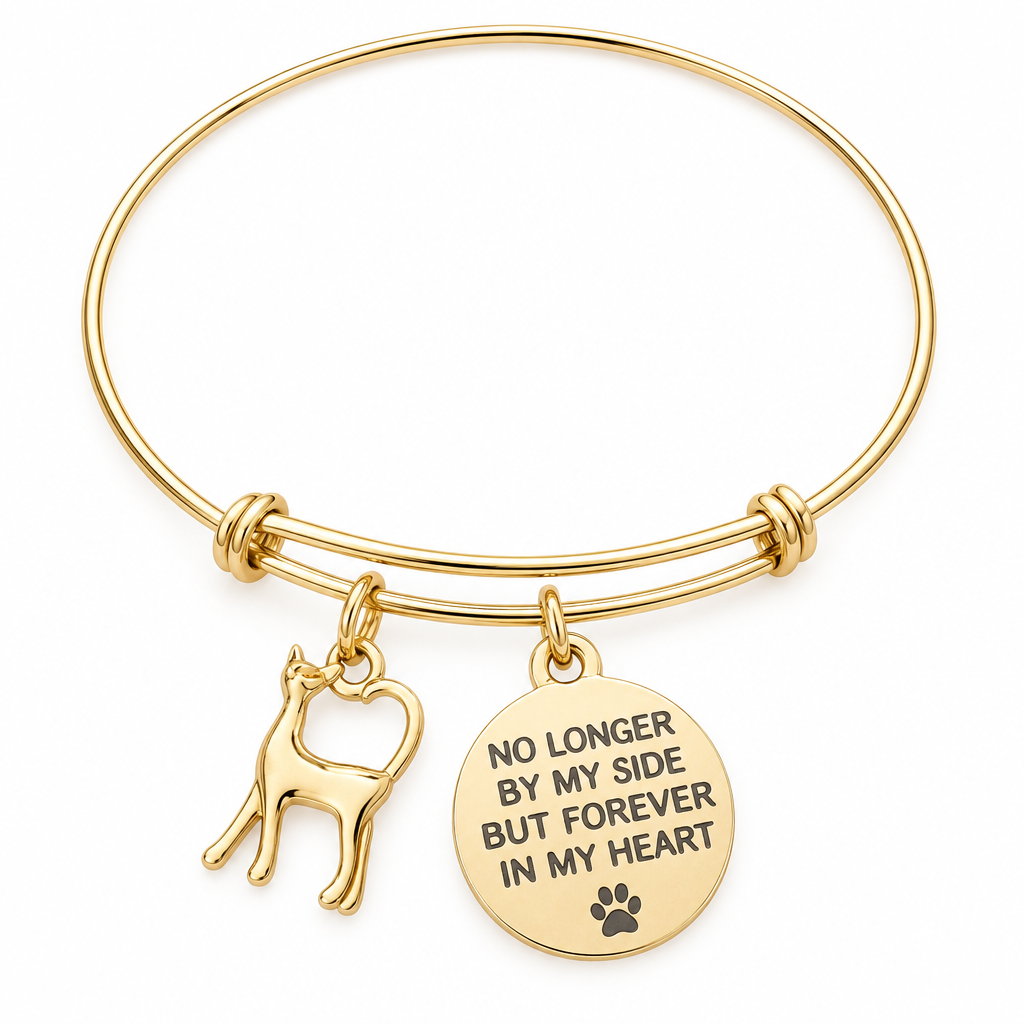 Gold bangle bracelet with a cat silhouette charm and a round disc charm engraved No Longer By My Side But Forever In My Heart with a paw print, shown on a warm-white studio background