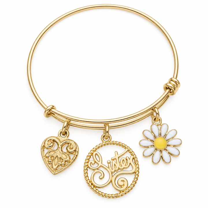 Gold bangle bracelet with a filigree heart charm, a round Sister script charm, and a white and yellow daisy charm, shown on a warm-white studio background