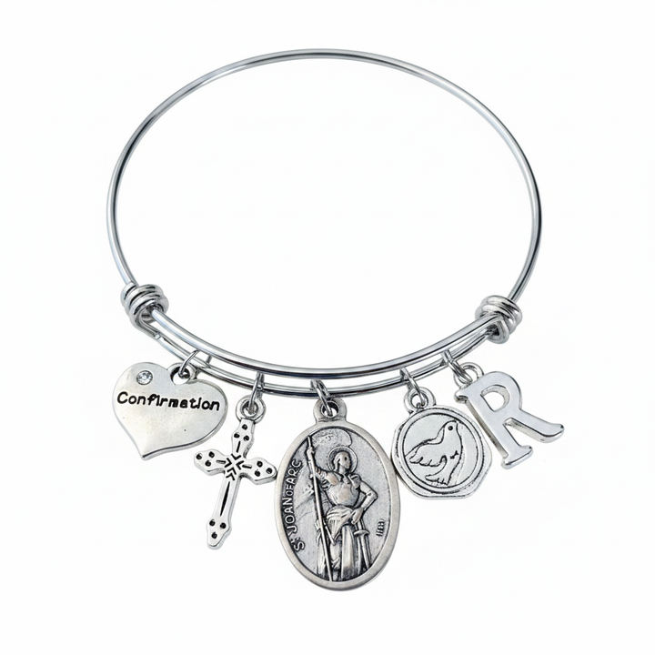 Saint Joan of Arc Confirmation charm bangle bracelet featuring St. Joan of Arc saint medal, cross charm, Confirmation heart, dove charm, and personalized initial charm in silver on white background