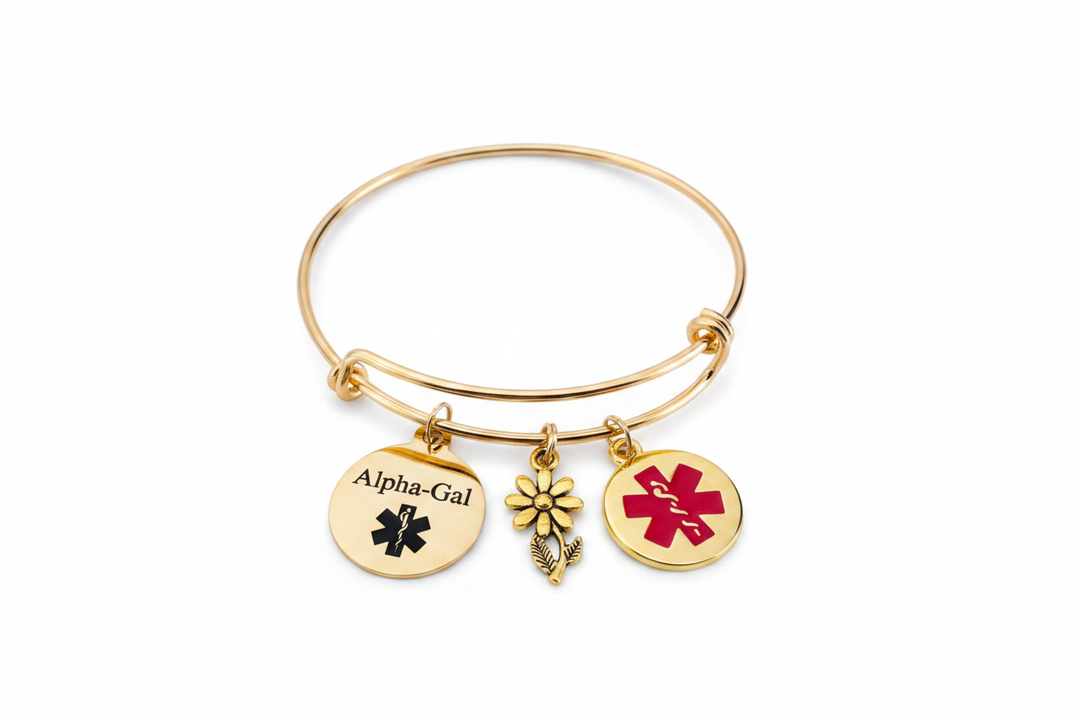 Gold expandable charm bangle bracelet with Alpha-Gal medical ID disc, red enamel medical alert disc, and gold daisy flower charm on white studio background
