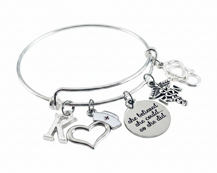 Silver expandable bangle bracelet with nurse-themed charms including K initial, open heart with nurse cap, RN caduceus, stethoscope, and inspirational disc charm reading she believed she could so she did, on white background