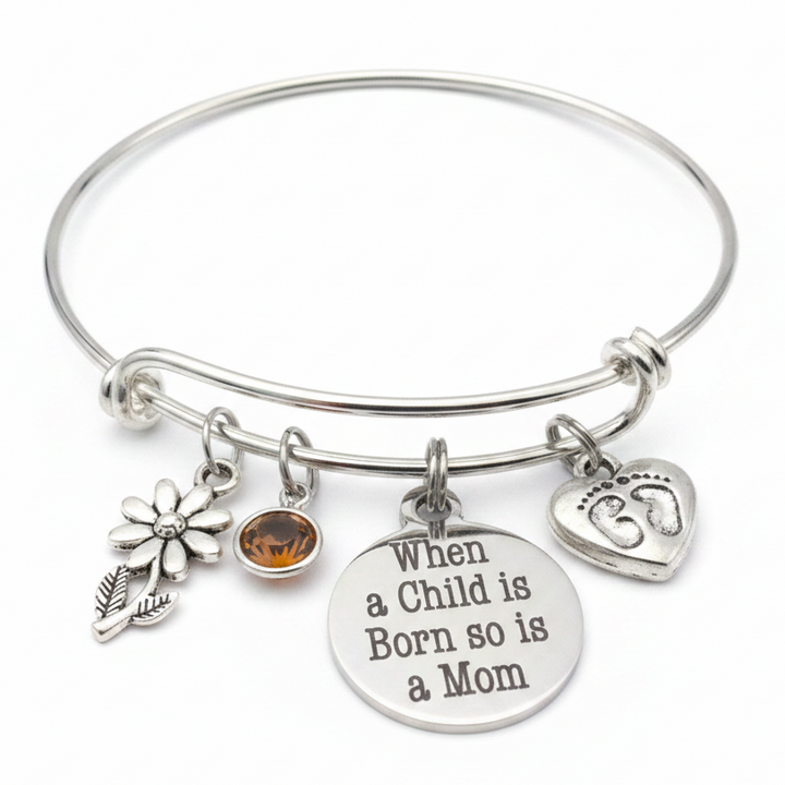 Silver expandable bangle bracelet with When a Child is Born So is a Mom disc charm, baby footprint heart charm, daisy flower charm, and amber birthstone crystal on white background – Jules Obsession new mom gift jewelry