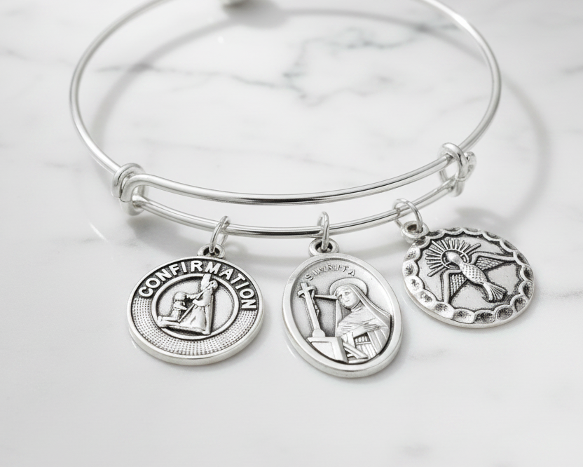 Silver adjustable bangle bracelet with three silver charms attached via jump rings: Confirmation charm, Saint Rita medal, and Holy Spirit dove charm