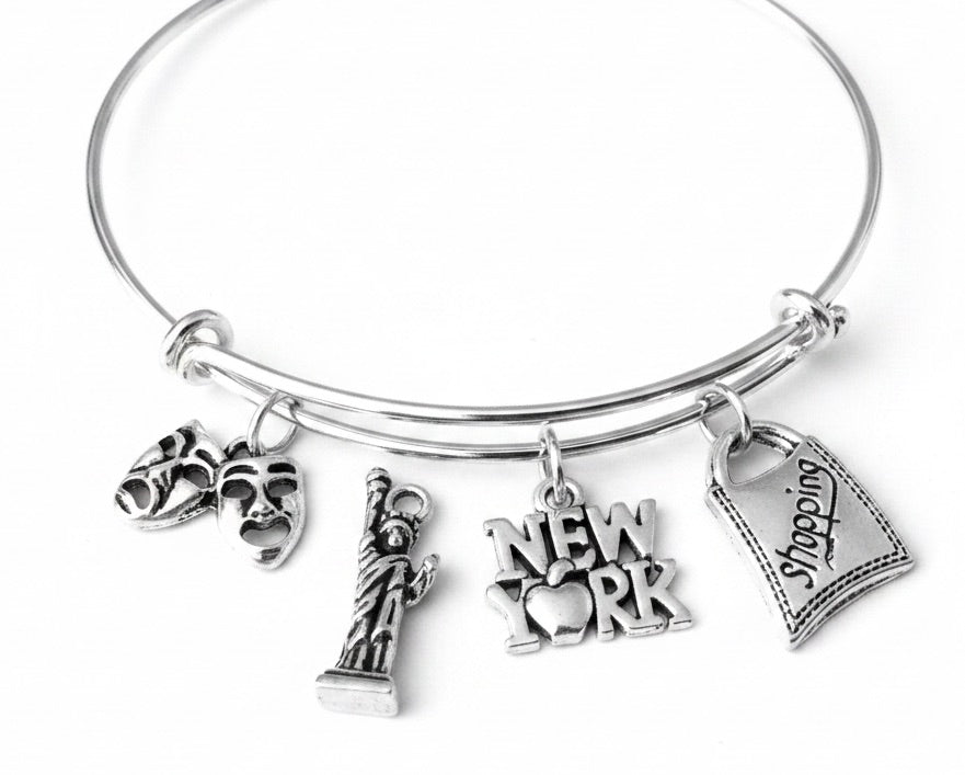 Silver expandable bangle bracelet with four New York City charms attached via jump rings: comedy and tragedy theater masks, Statue of Liberty, "New York" text charm with heart, and a shopping bag charm engraved with "Shopping" — displayed on a white background.
