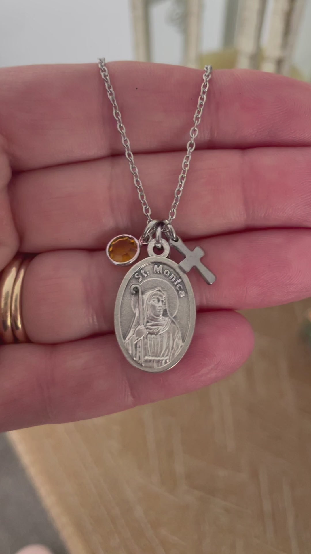 Oxidized silver Saint Monica medal necklace on wood surface showing St. Monica oval medal with engraved portrait, silver cross charm, and amber golden crystal birthstone charm on a stainless steel cable chain