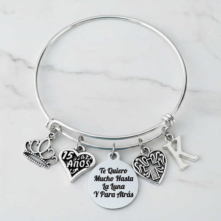 Silver expandable bangle bracelet with crown charm, 15 Años heart charm, Te Quiero Mucho Hasta La Luna engraved disc, filigree cross charm, and personalized initial charm — meaningful Quinceañera gift for girls