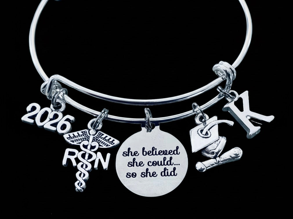 Silver expandable bangle bracelet with 2026 year charm, graduation cap and diploma charm, RN caduceus medical symbol charm, stethoscope charm, and 'She Believed She Could So She Did' engraved disc — personalized 2026 RN nurse graduation gift bracelet by Jules Obsession